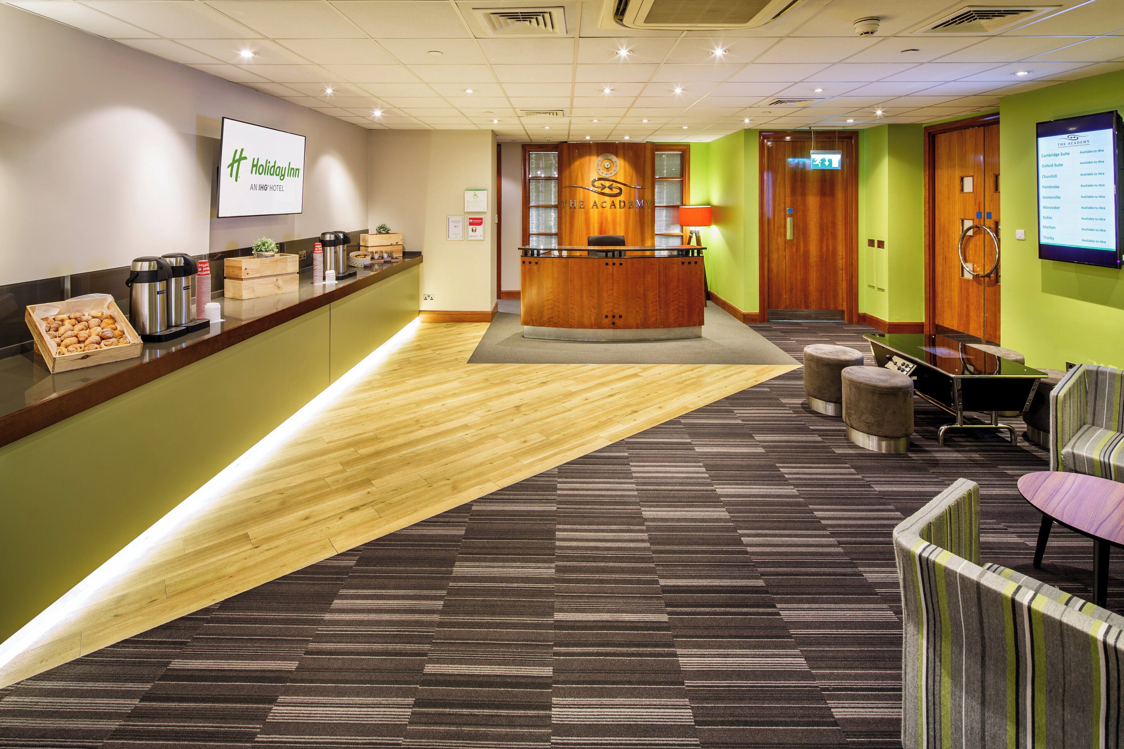 A hotel reception area with a wooden reception desk, a digital screen displaying room availability, and a carpeted floor.