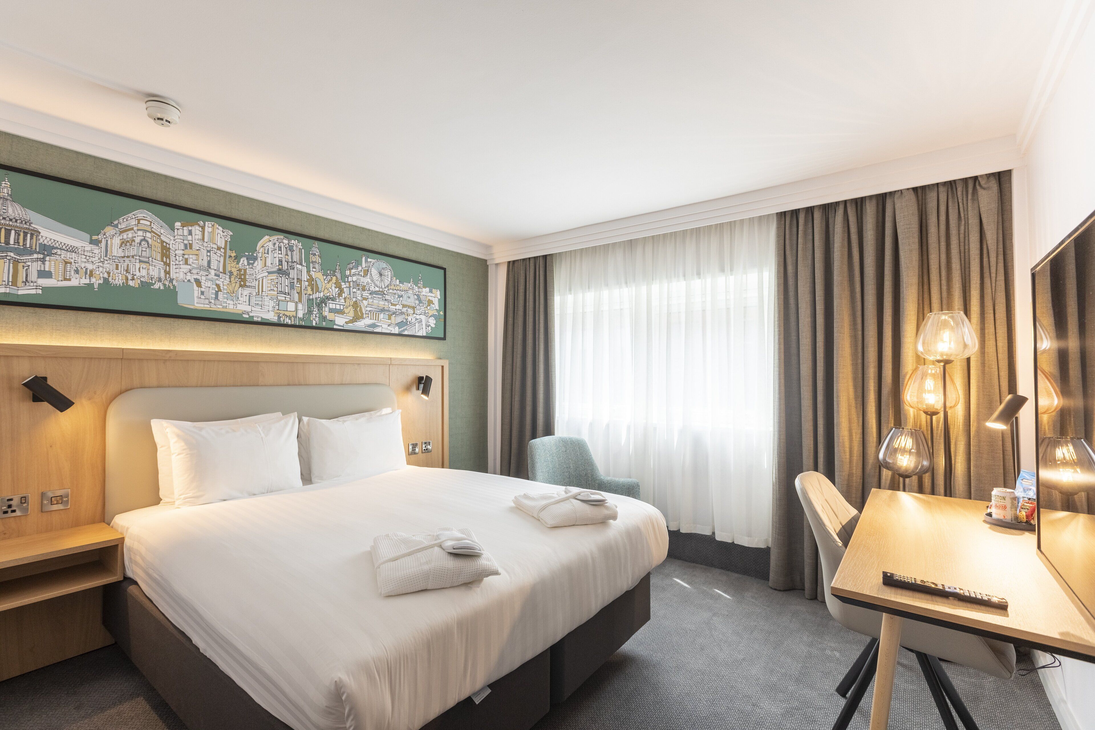 Camera d'albergo con un letto grande, una scrivania, una sedia, un quadro appeso al muro e una finestra con tende.