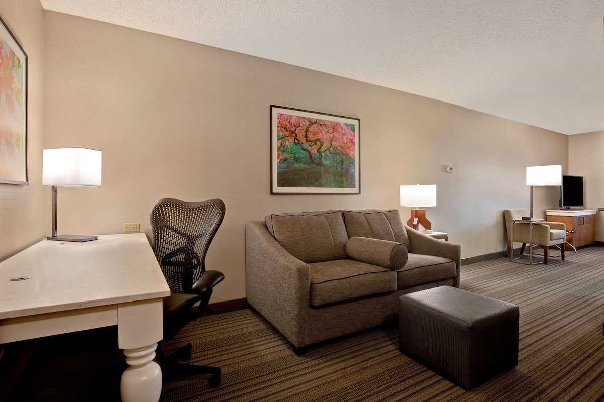 A hotel room with a sofa, ottoman, chair, table, lamp, TV, and artwork on the wall.