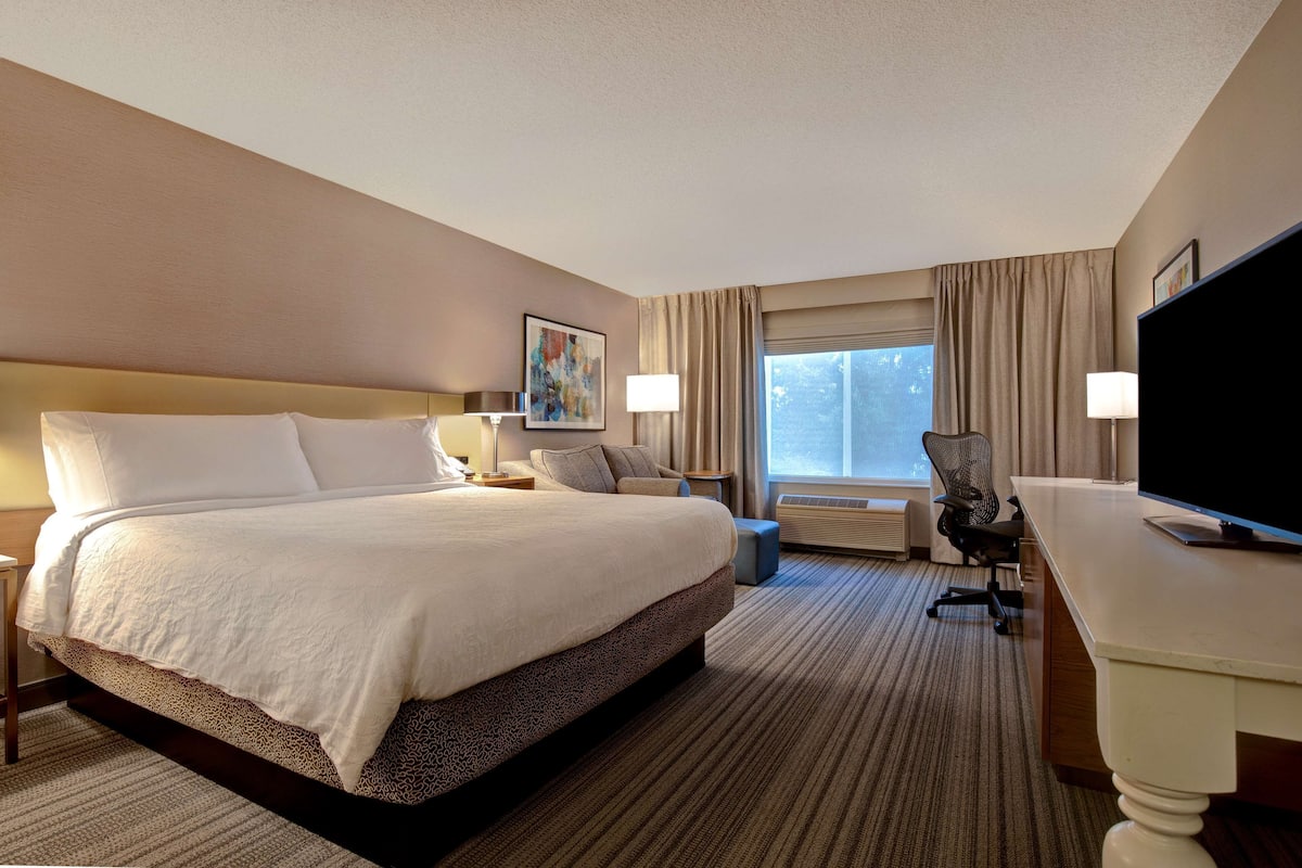 A hotel room with a large bed, a desk, a chair, a TV, and a window with curtains.