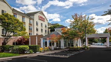 A hotel with a covered entrance, a parking lot, and a building with a sign.