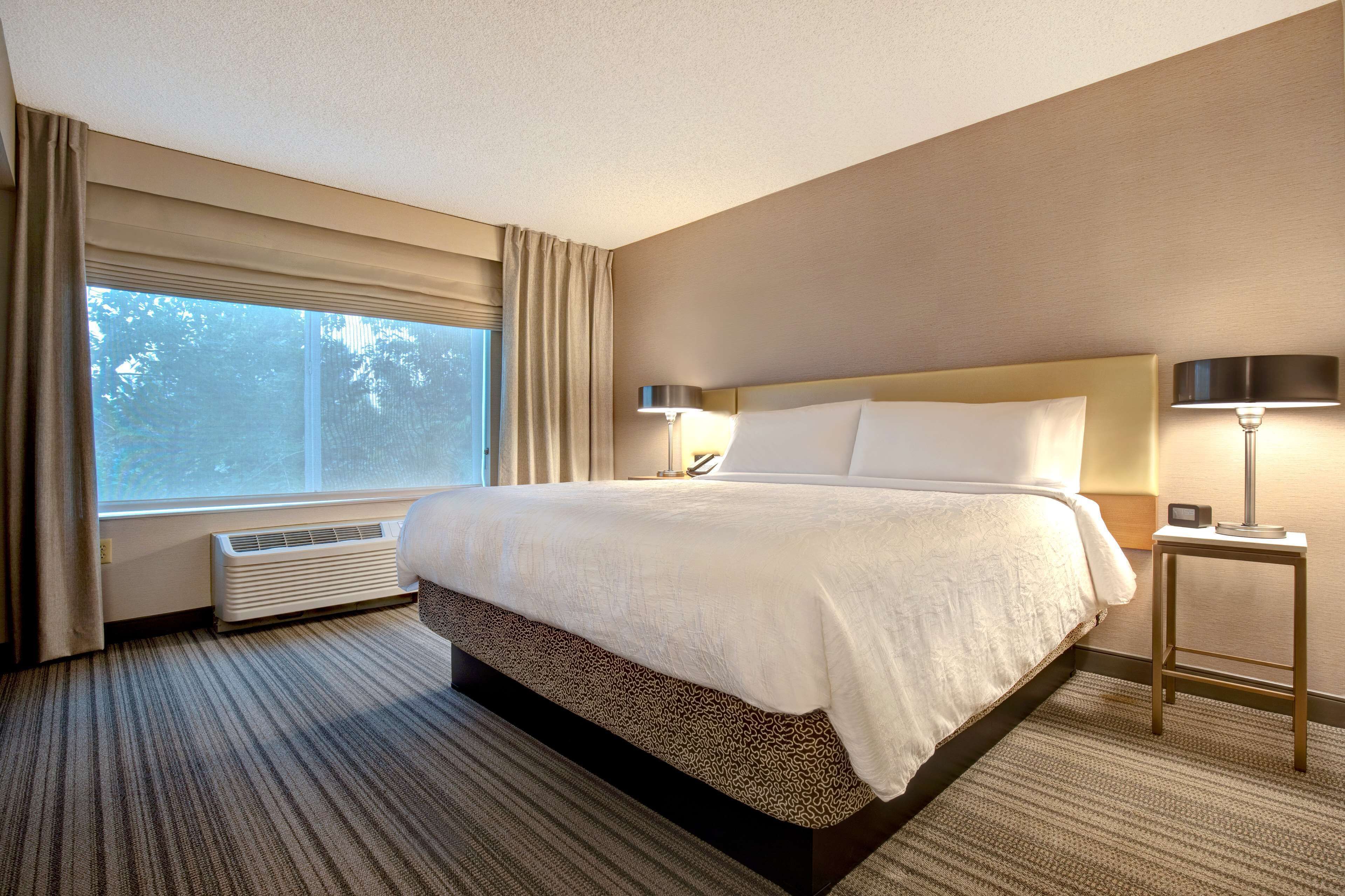 Habitación de hotel con una cama grande, una mesita de noche con lámpara, una ventana con cortinas y un calentador fijado a la pared.