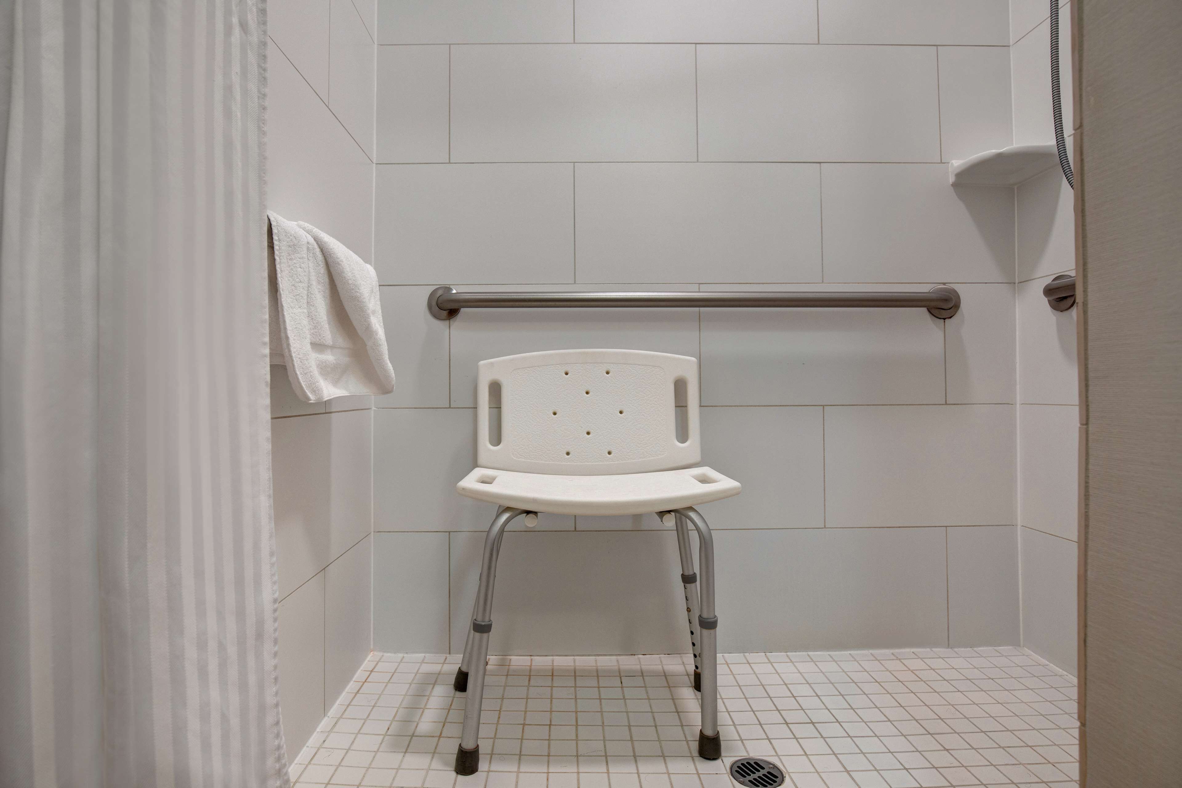 A white wheelchair-accessible shower stall with a grab bar, a white towel hanging on the wall, and a tiled floor.