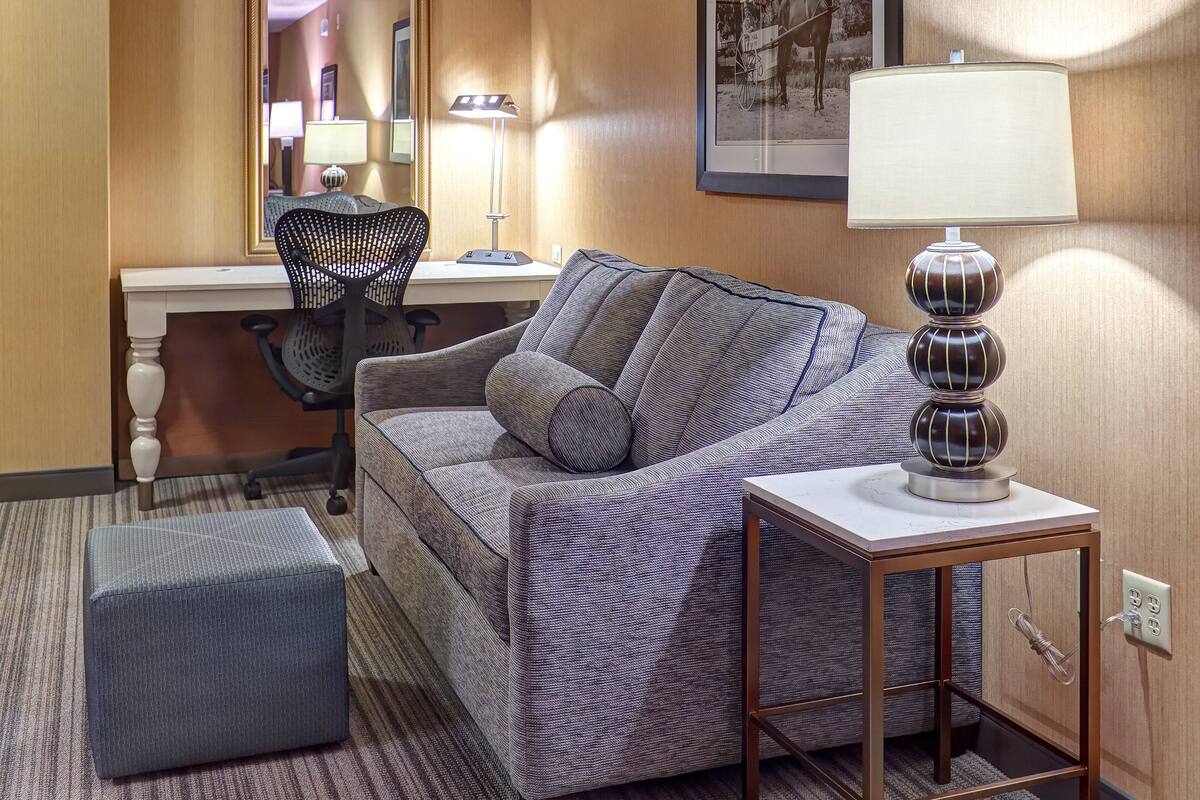 A hotel room with a grey sofa, a small table with a lamp, a chair, a desk, and a framed picture on the wall.