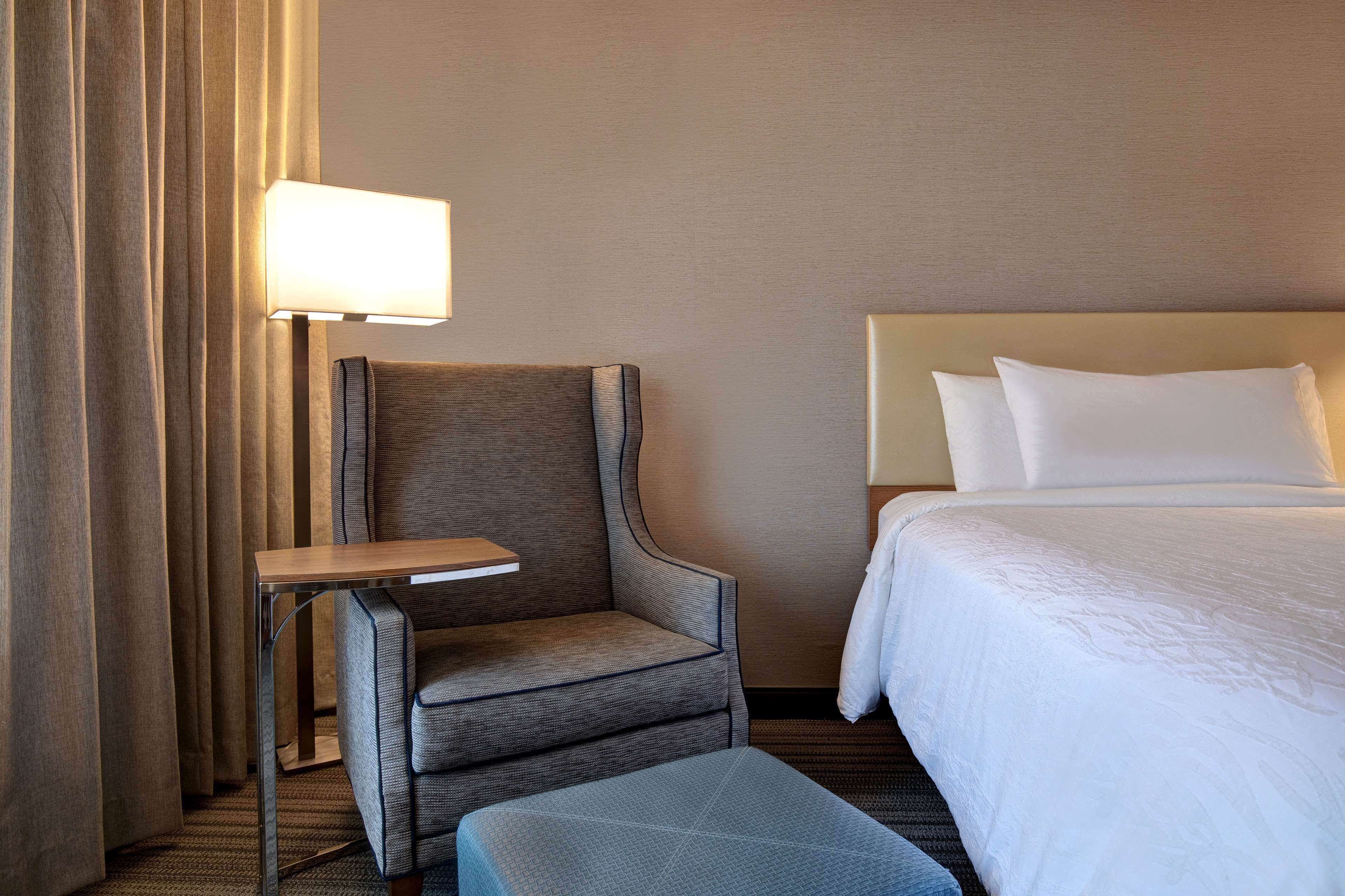 A hotel room with a bed, a chair, a small table, and a lamp.