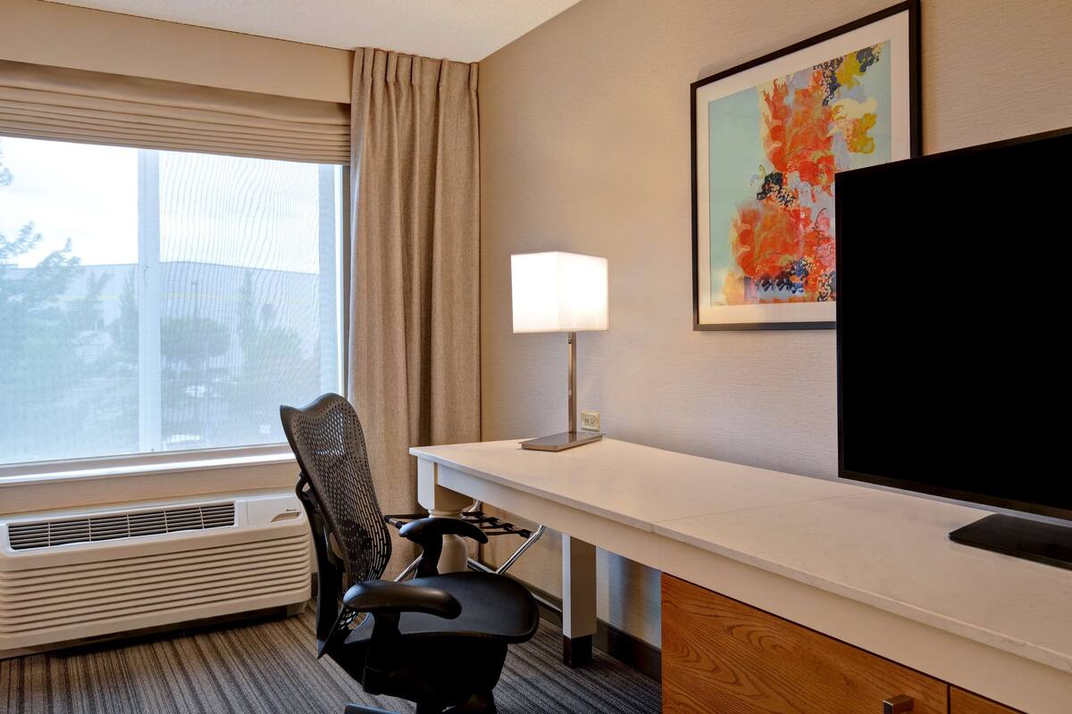 A hotel room with a desk, chair, air conditioning unit, window with curtains, and a large flat-screen TV.