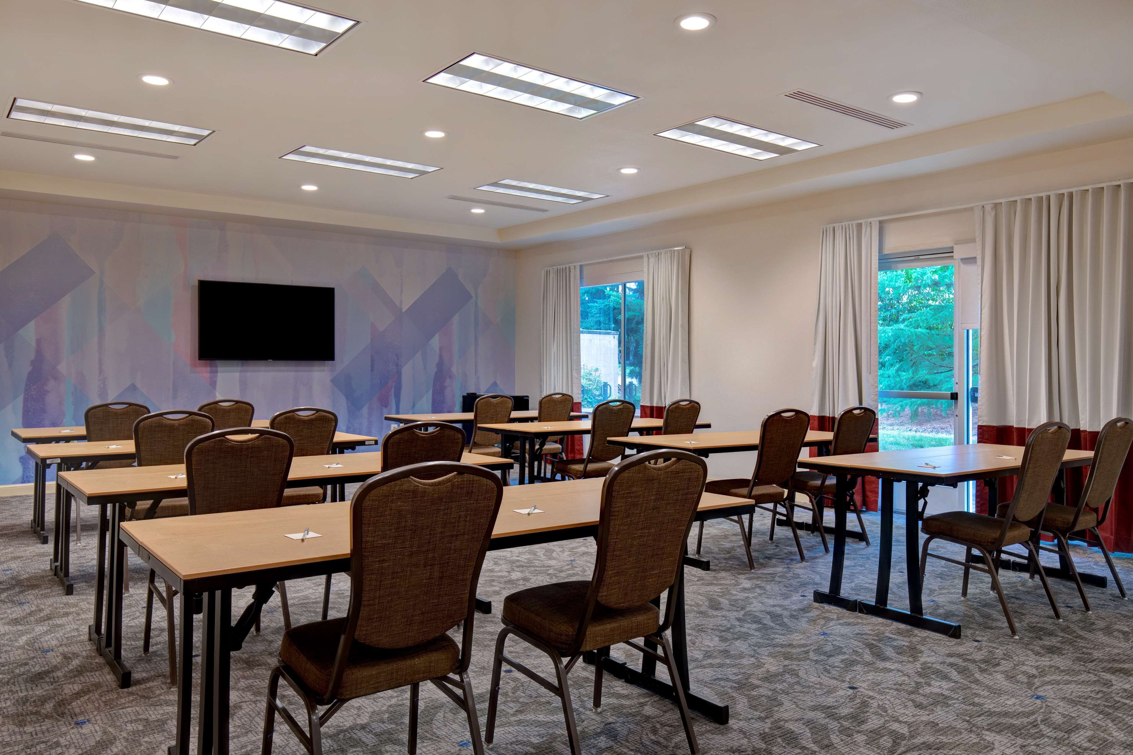 A conference room with multiple tables and chairs, a flat-screen TV mounted on the wall, and large windows with curtains.