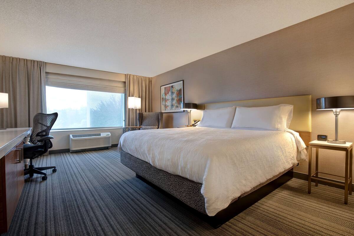 A hotel room with a large bed, a desk with a chair, a standing lamp, and a window with curtains.