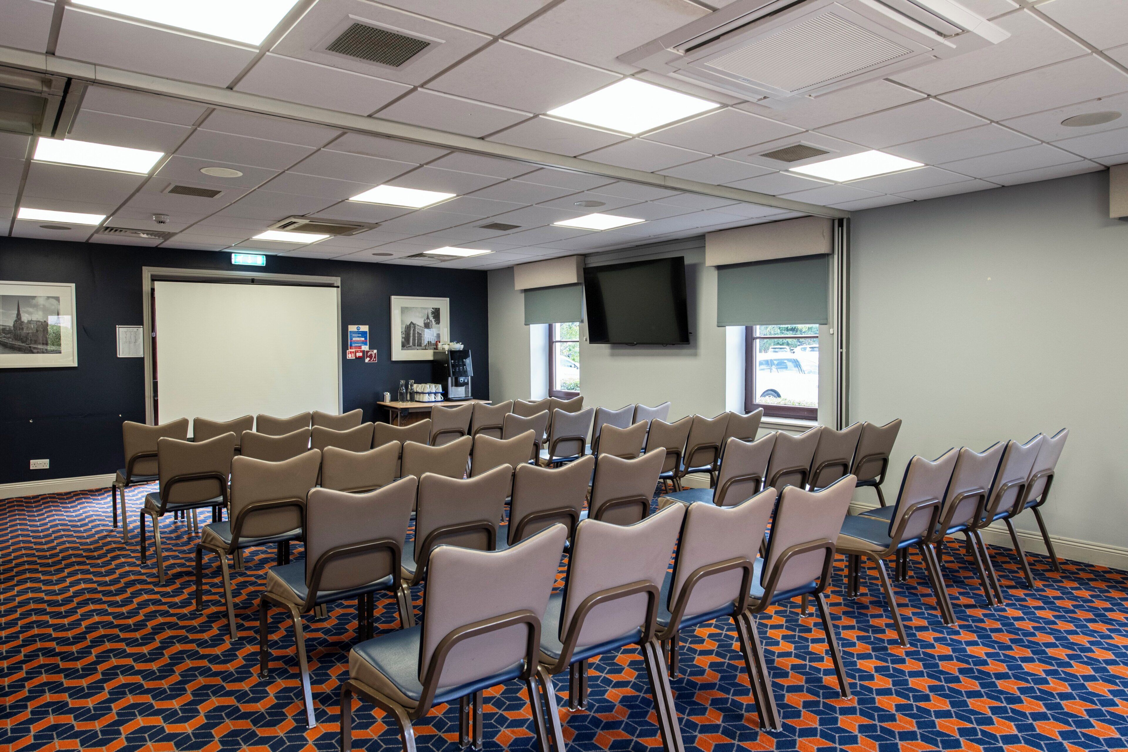 A conference room with rows of chairs, a projector screen, a television, and a patterned carpet.