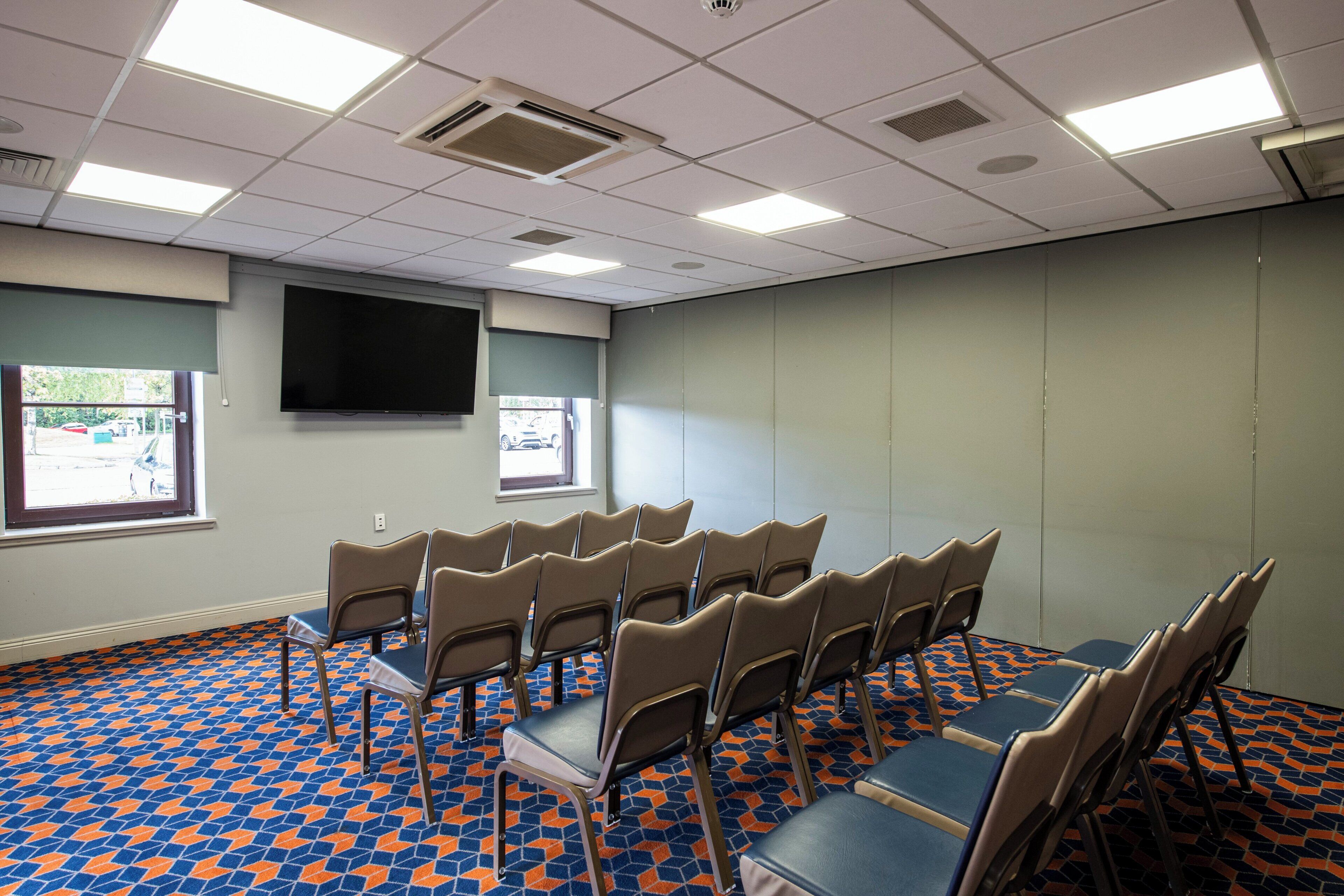 A conference room with a patterned floor, rows of chairs, a large screen, and a window with a view of a parking lot.