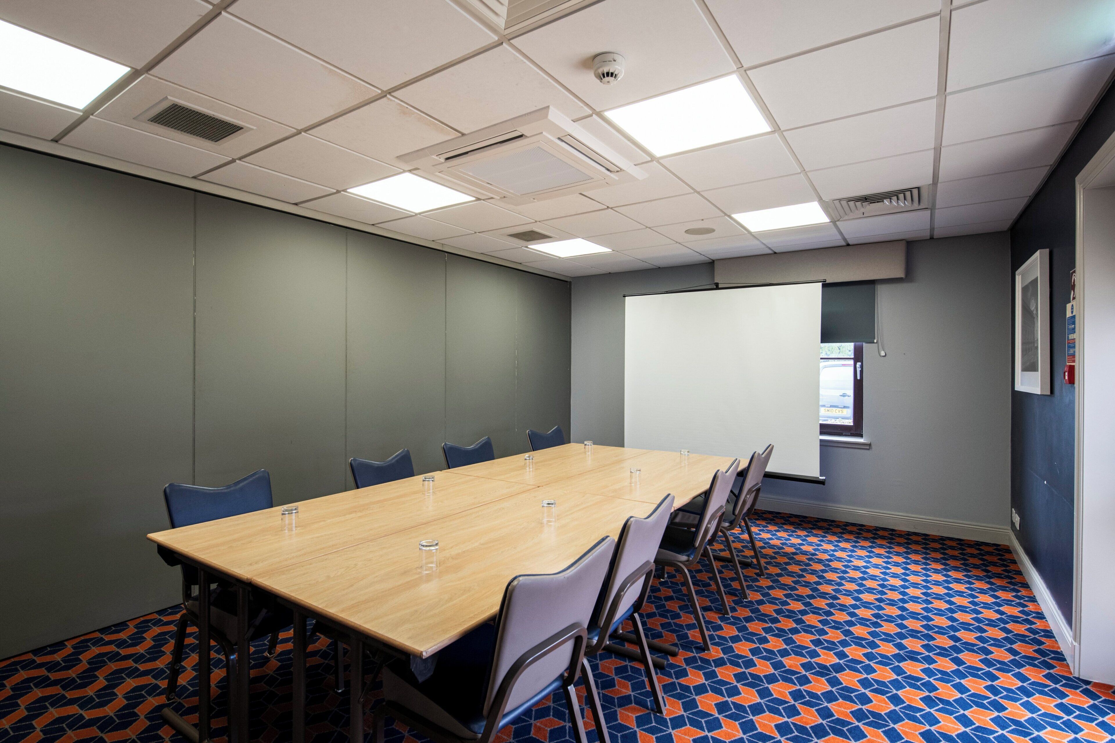 A conference room with a long table, chairs, a projector screen, and patterned carpet.