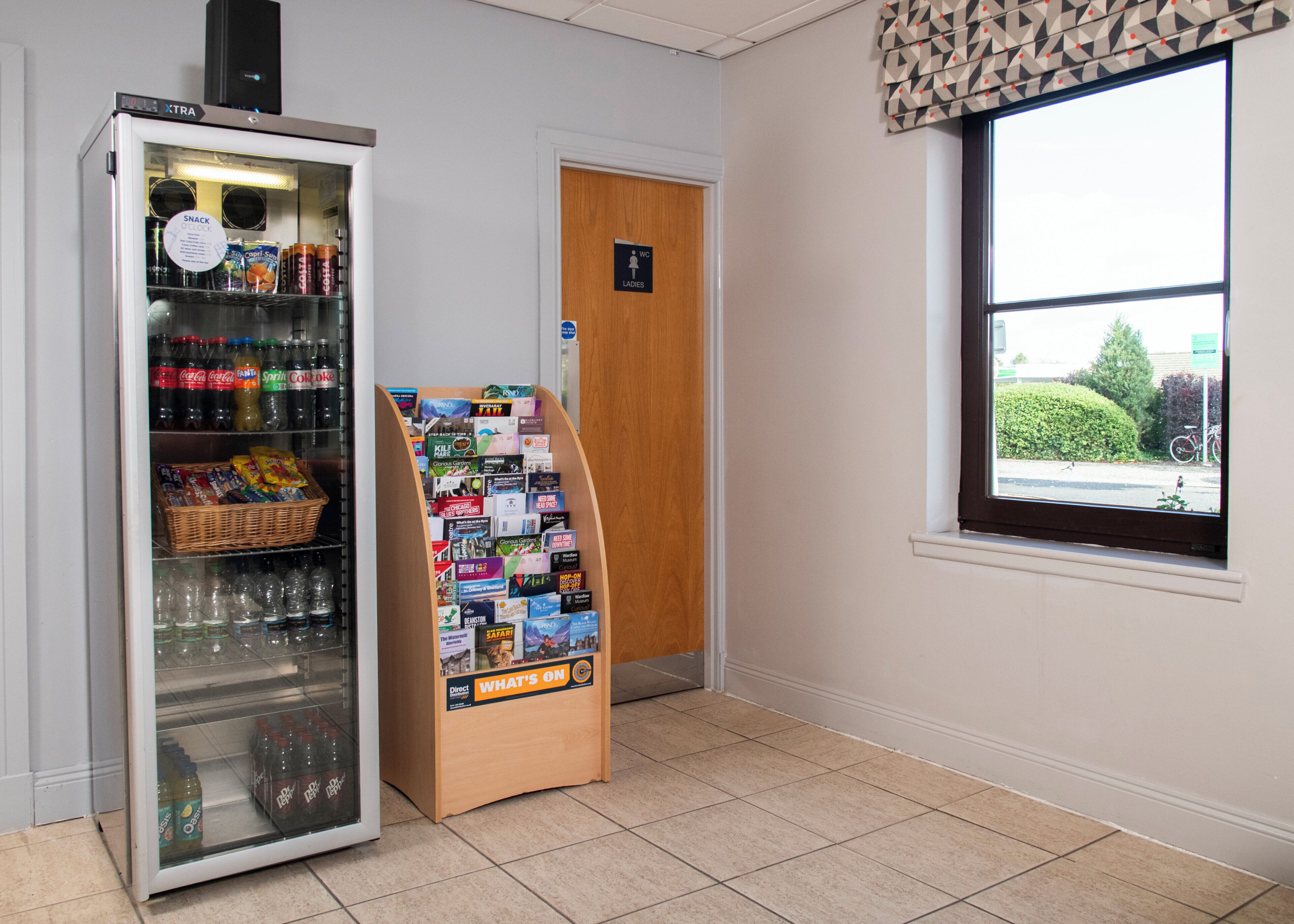 A vending machine stocked with beverages and snacks, a bulletin board with flyers, and a women's restroom door.