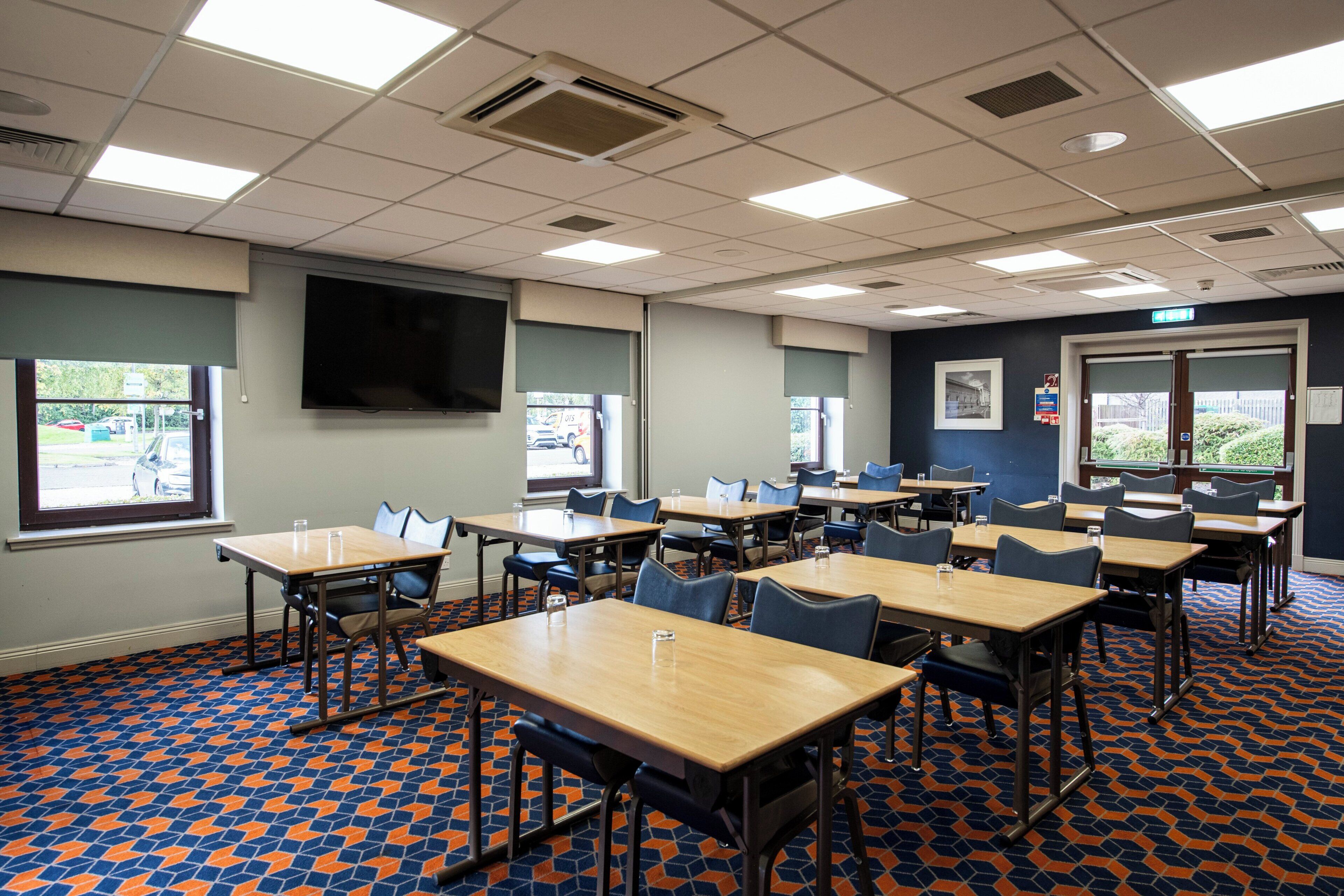Une salle de classe avec des tables et des chaises, un grand écran, des fenêtres et un tapis à motifs.
