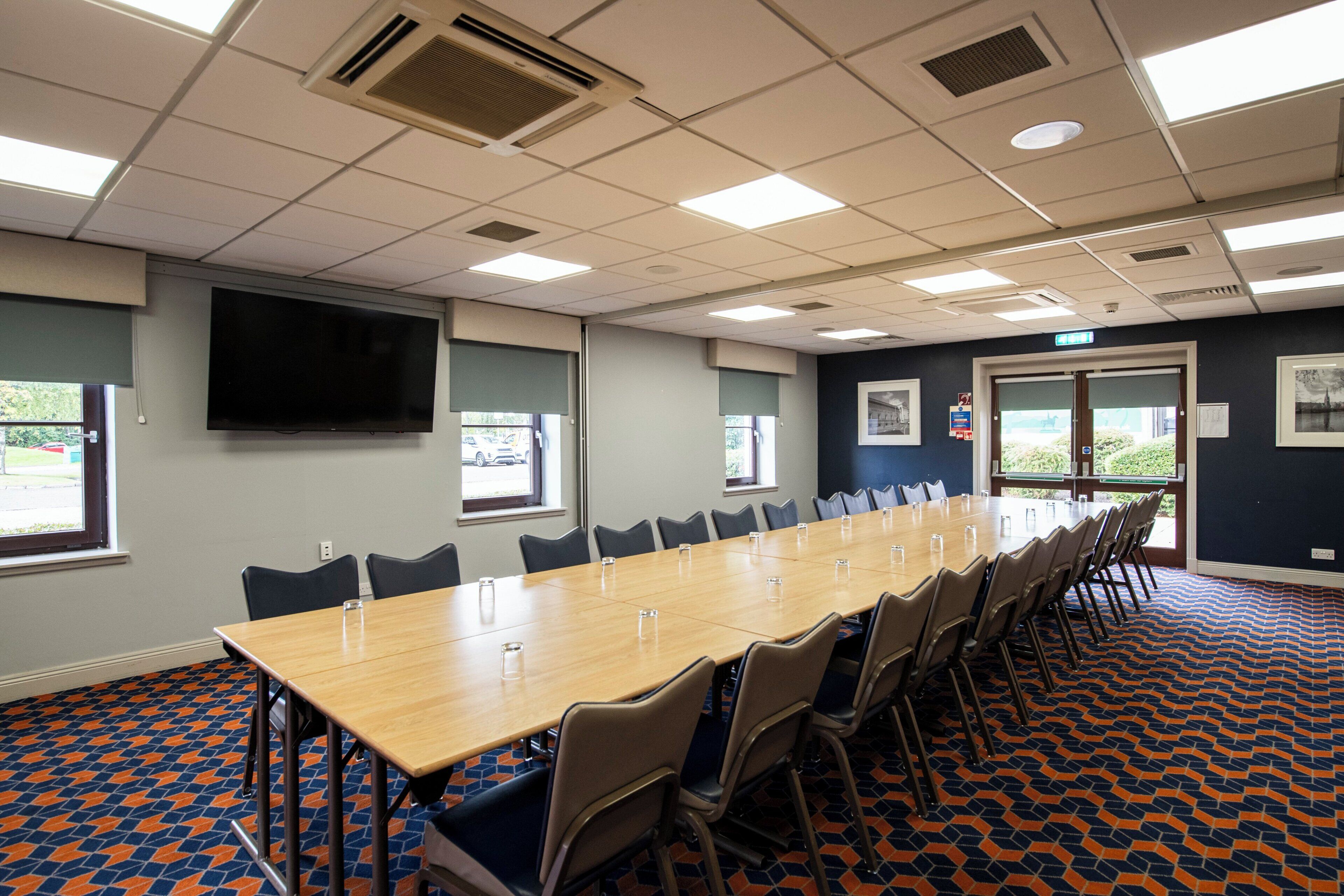 A conference room with a long table, chairs, a flat-screen TV, windows, and patterned carpet.