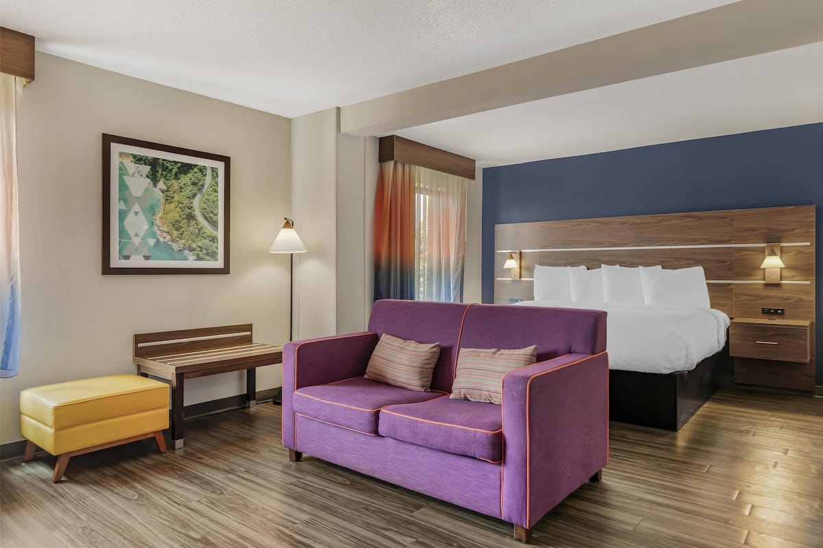 A hotel room with a purple sofa, a wooden bench, a small yellow ottoman, a bed with white linens, a nightstand, and a wall-mounted lamp.