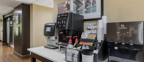 A hotel reception area with a coffee machine, self-serve coffee station, and a framed picture of a bridge.