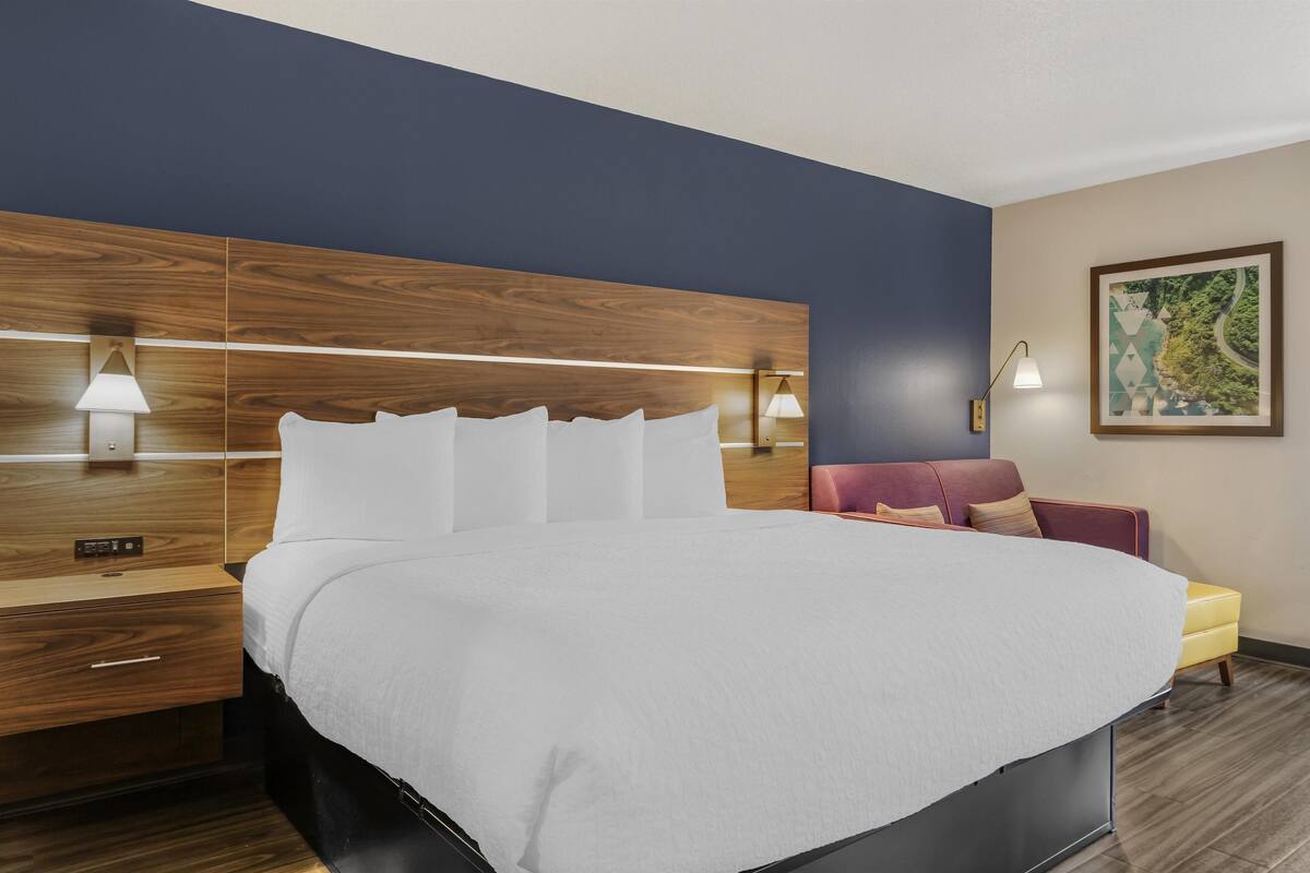 A hotel room with a large bed, wooden headboard, bedside lamps, a small table, a chair, and a framed picture on the wall.