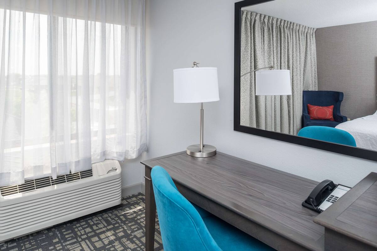 A hotel room with a desk, a chair, a telephone, a mirror, and a window with sheer curtains.