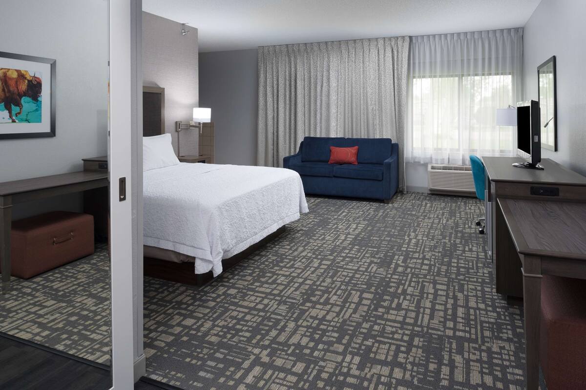 A hotel room with a bed, a blue sofa, a desk with a computer, and a window with curtains.