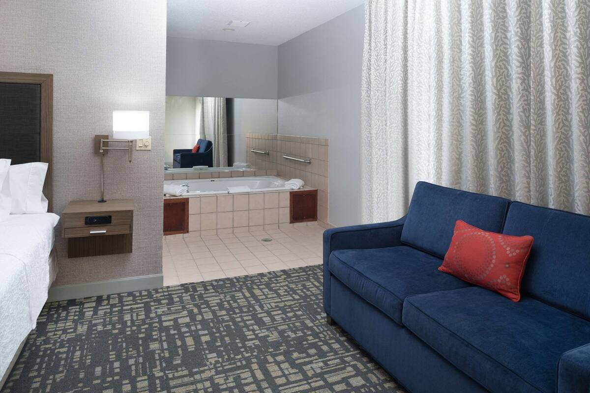 A hotel room with a bed, a blue sofa, a bathroom with a bathtub, and a patterned rug.