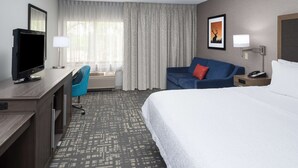A hotel room with a bed, a desk with a television, a blue chair, a blue sofa with a red pillow, and a framed picture on the wall. - Hampton Inn North Sioux City (North Sioux City)