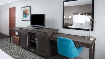 Ein Hotelzimmer mit Schreibtisch, Fernseher, Spiegel, blauem Sessel, braunem Koffer und einem gemusterten Teppich.