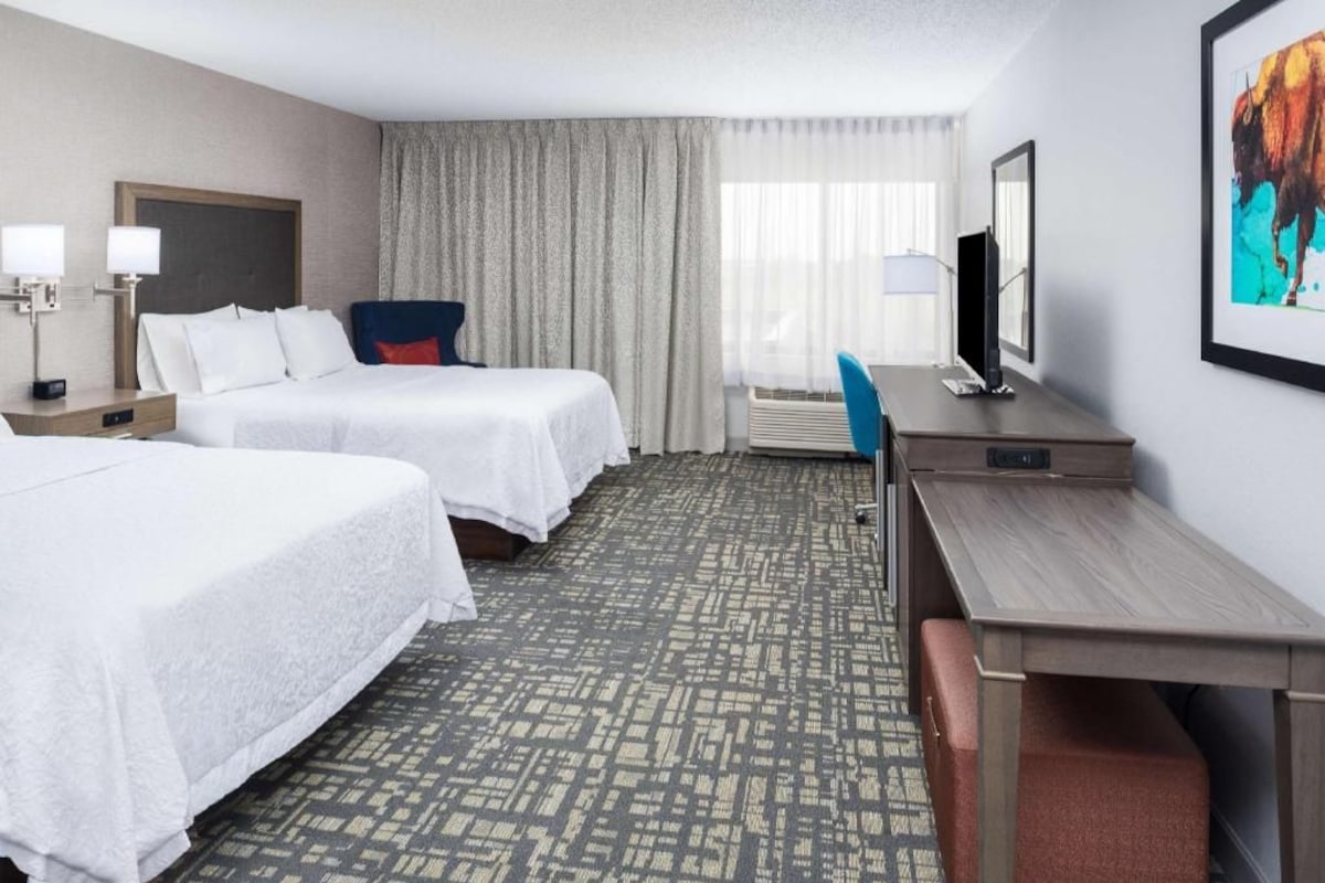 A hotel room with two beds, a desk, a chair, a TV, and a large window with curtains.