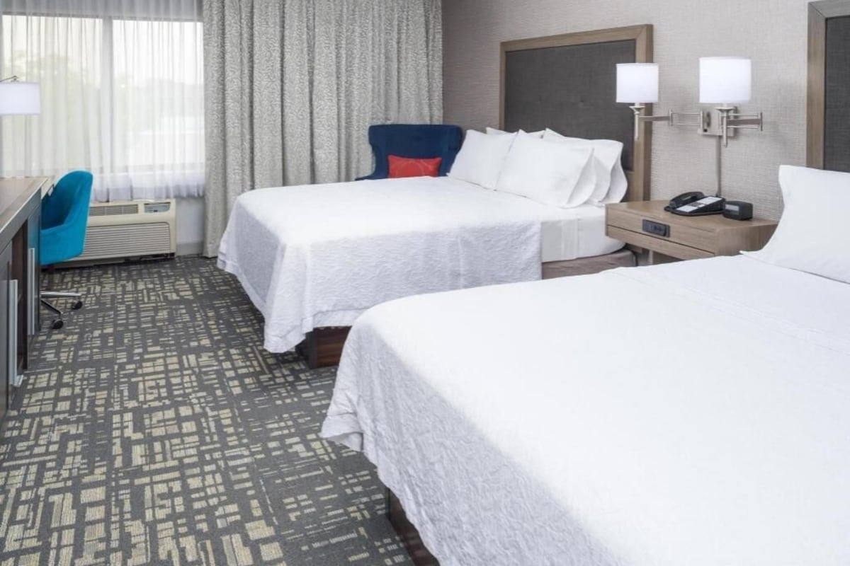 A hotel room with two beds, a desk, a chair, a nightstand, and a lamp.