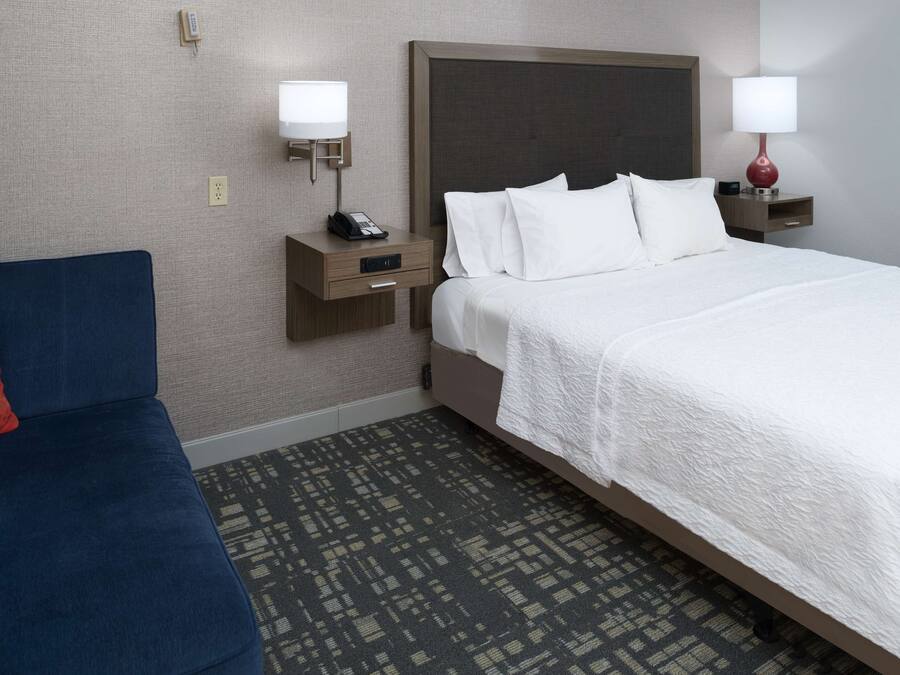 Una habitación de hotel con cama, un sofá azul, una mesita de noche con lámpara y un enchufe de pared.