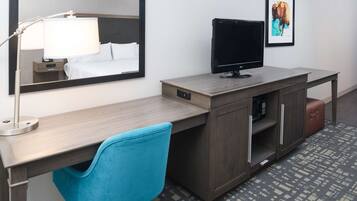 Habitación de hotel con un escritorio de madera, una silla azul, un televisor de pantalla plana, una lámpara de noche, una cama con ropa de cama blanca, un cuadro en la pared y una alfombra estampada.
