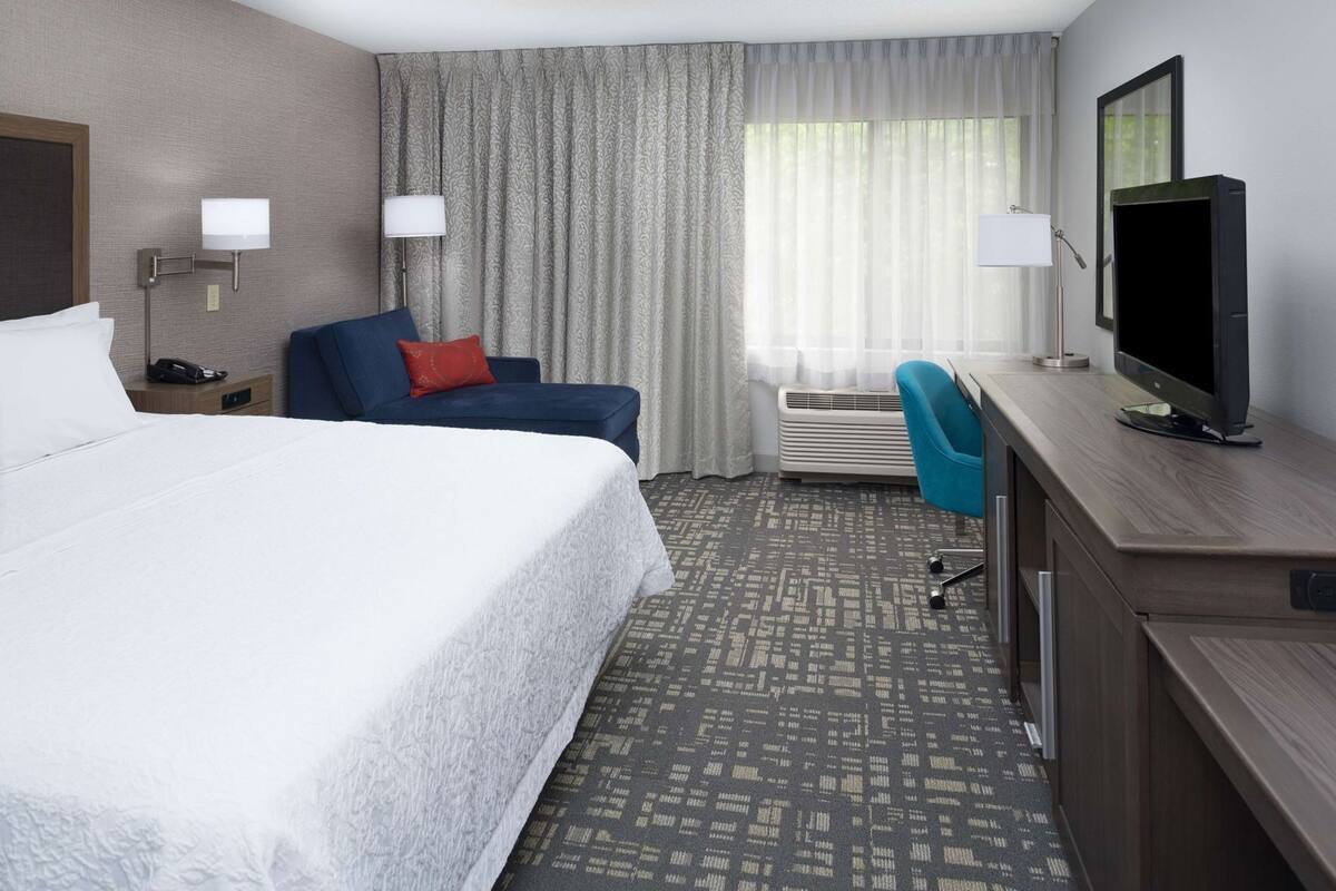 A hotel room with a large bed, a desk with a flat-screen TV, a chair, a desk lamp, a telephone, and a window with curtains.