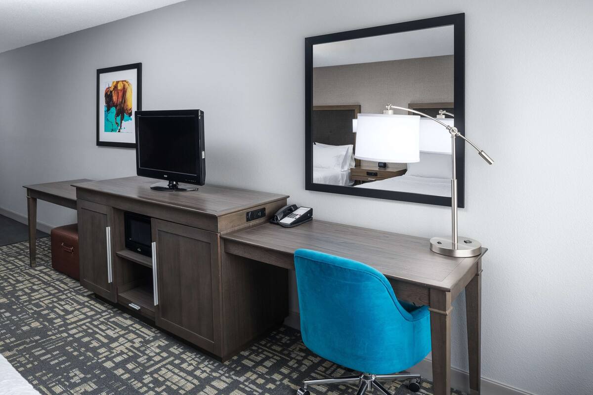 A hotel room with a wooden desk, a blue chair, a flat-screen TV, a lamp, a telephone, and a framed picture on the wall.