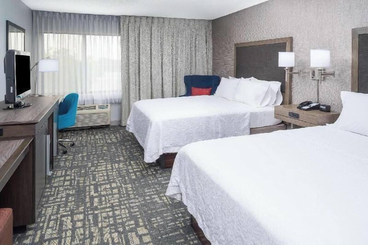 A hotel room with two beds, a desk, a chair, a TV, and a lamp.