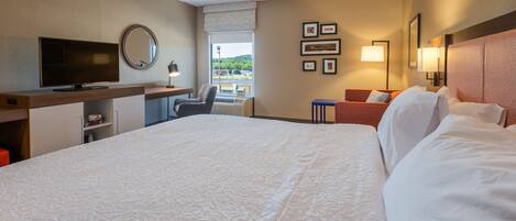 A hotel room with a large bed, a desk with a flat-screen TV, a chair, a small table, and a window with a view of the outdoors.