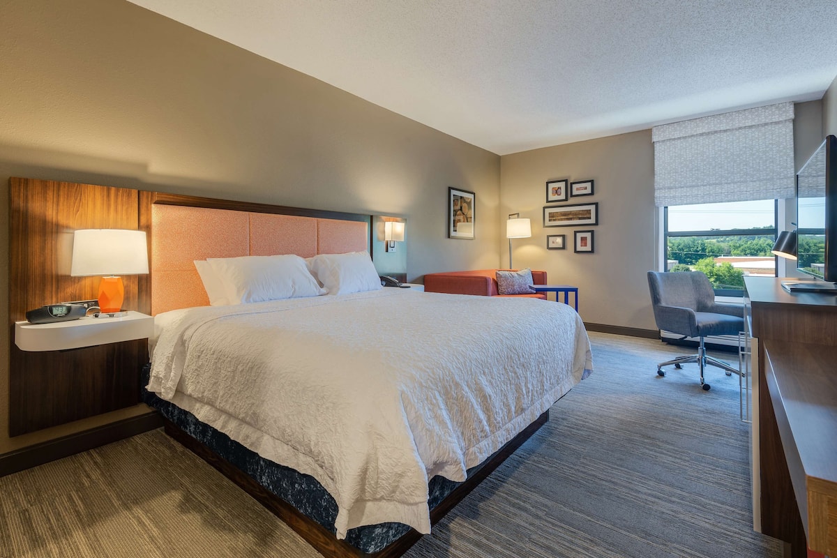 A hotel room with a large bed, a desk, a chair, a nightstand, and a window with a view.