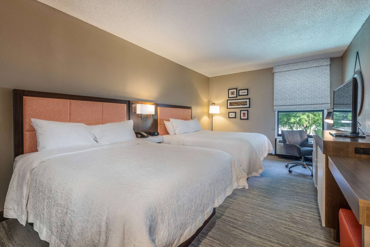 A hotel room with two beds, a desk, a chair, a TV, and framed pictures on the wall.