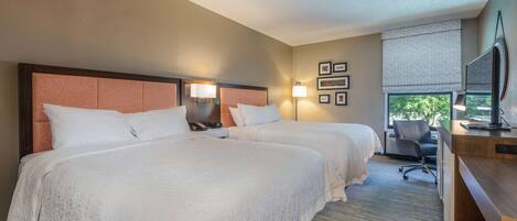 A hotel room with two beds, a desk, a chair, a TV, and framed pictures on the wall.