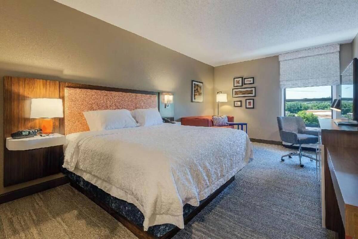 A hotel room with a large bed, a desk, a chair, a desk lamp, a nightstand, and a window with a view.