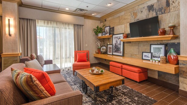 A modern living room with a sofa, coffee table, and red ottoman. There is a TV mounted on the wall, a fireplace, and a window with curtains. - Hampton Inn Wilkesboro (Wilkesboro)