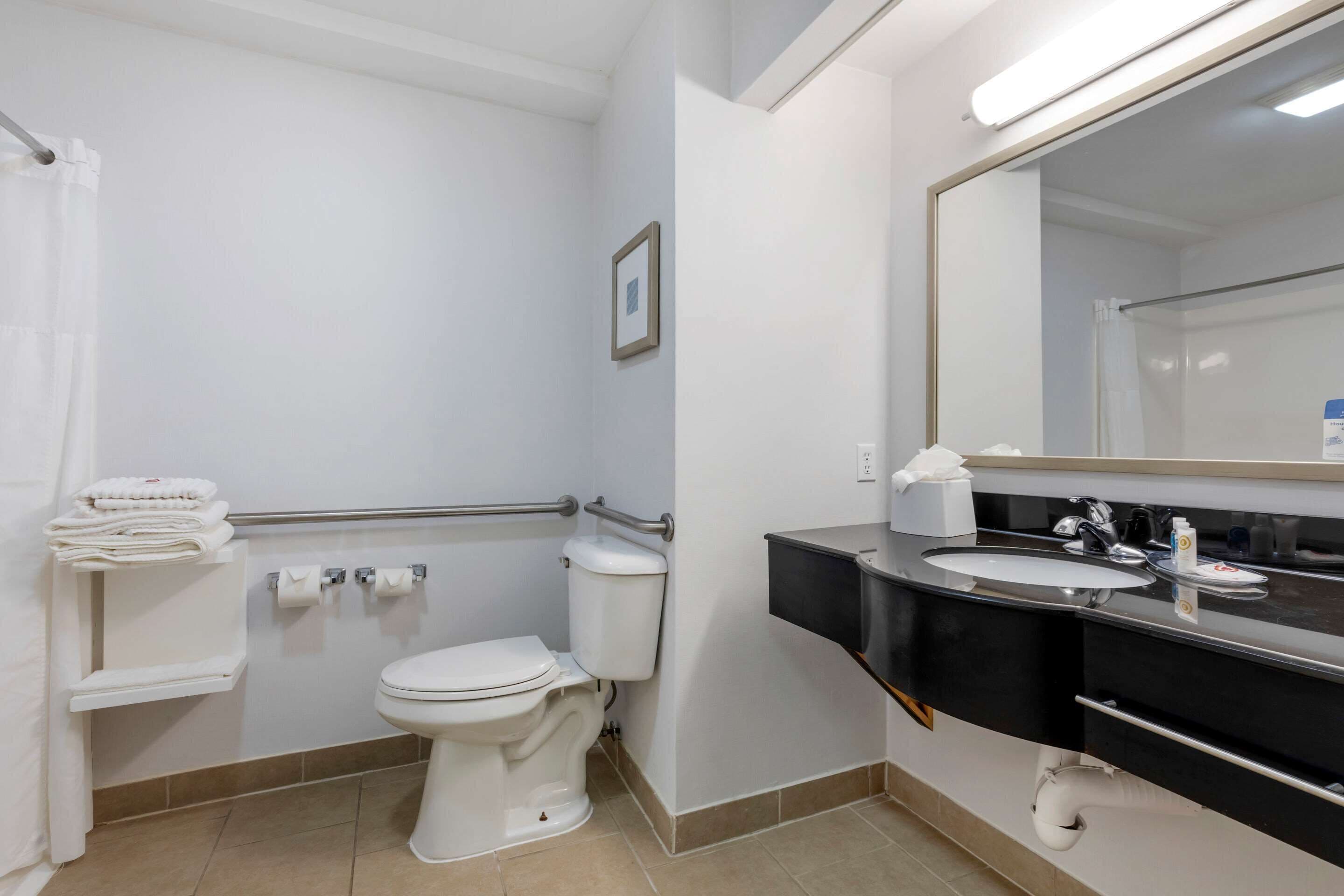 Une salle de bain avec des toilettes blanches, un lavabo et un miroir. On y trouve un porte-serviettes avec des serviettes pliées, un tableau accroché au mur et une barre d’appui.