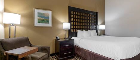 A hotel room with a bed, a desk, a chair, a nightstand, and a wall-mounted lamp.