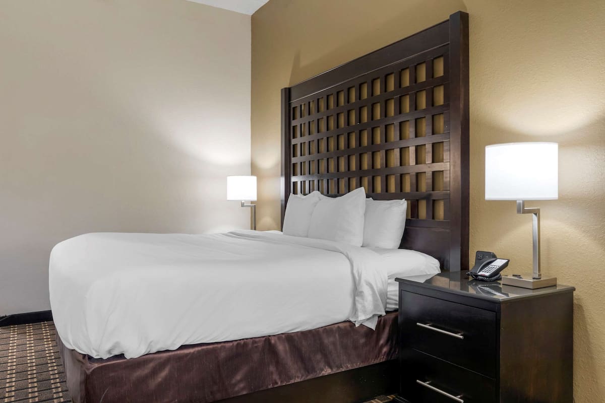 A hotel room with a large bed, a wooden headboard, two bedside lamps, a telephone, and a small desk with drawers.