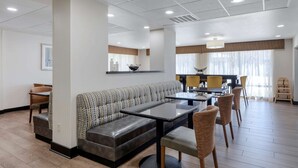 A modern hotel lobby with a seating area, a dining table, and a kitchen in the background.
