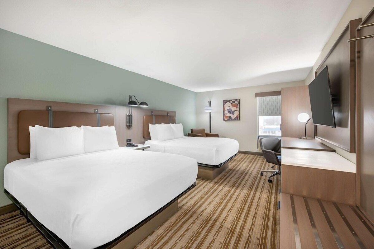 A hotel room with two beds, a desk, a TV, and a window.