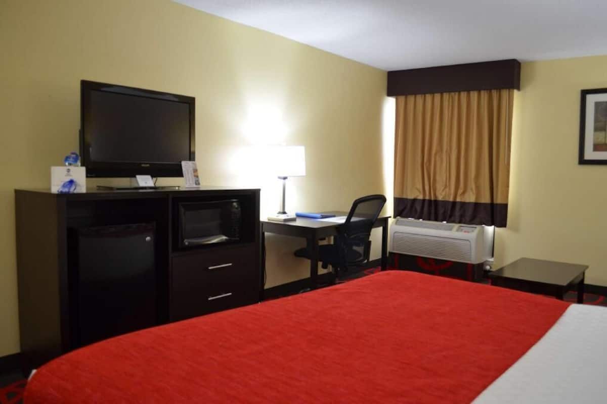 A hotel room with a bed, a desk with a computer, a television, a mini-fridge, and an air conditioning unit.