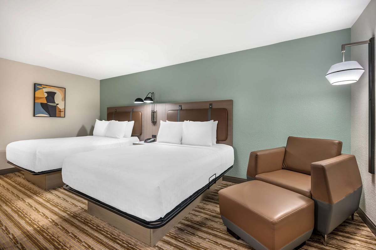 A hotel room with two beds, a brown leather armchair, a lamp, and a painting on the wall.