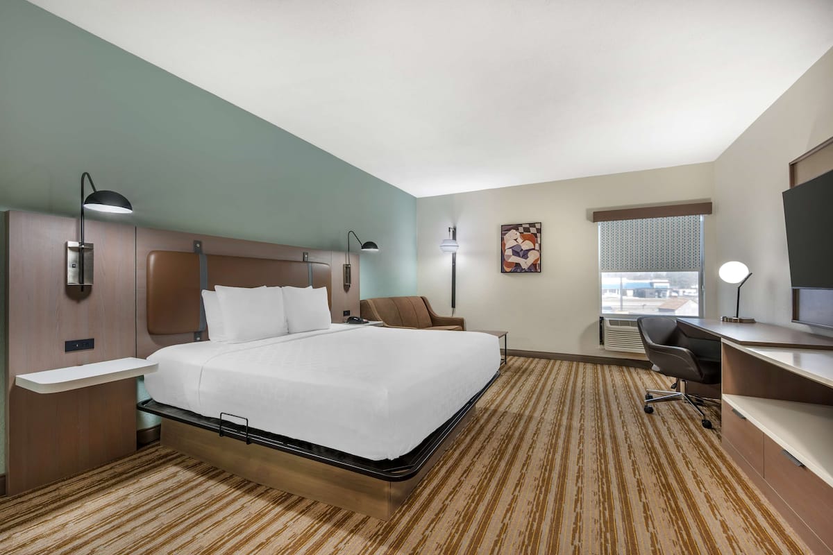 A hotel room with a large bed, a desk with a chair, a TV, a window with blinds, and a painting on the wall.