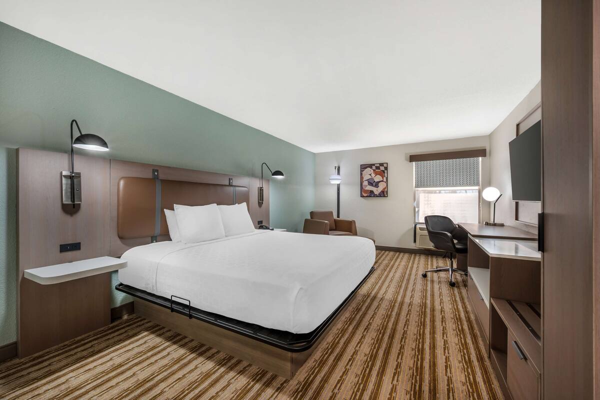 A hotel room with a large bed, bedside tables, a desk, a chair, and a window with blinds.