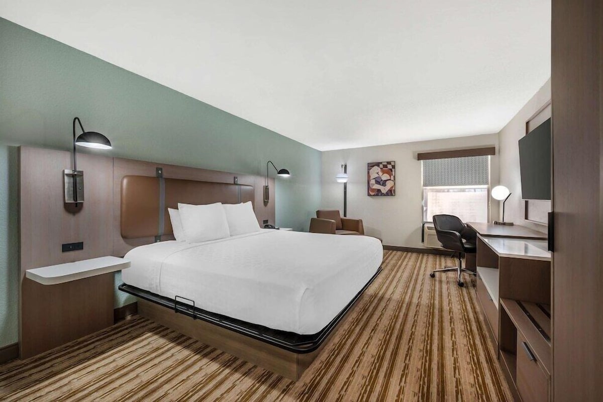 A hotel room with a bed, bedside tables, a desk, a chair, a desk lamp, a wall-mounted TV, and a window with blinds.