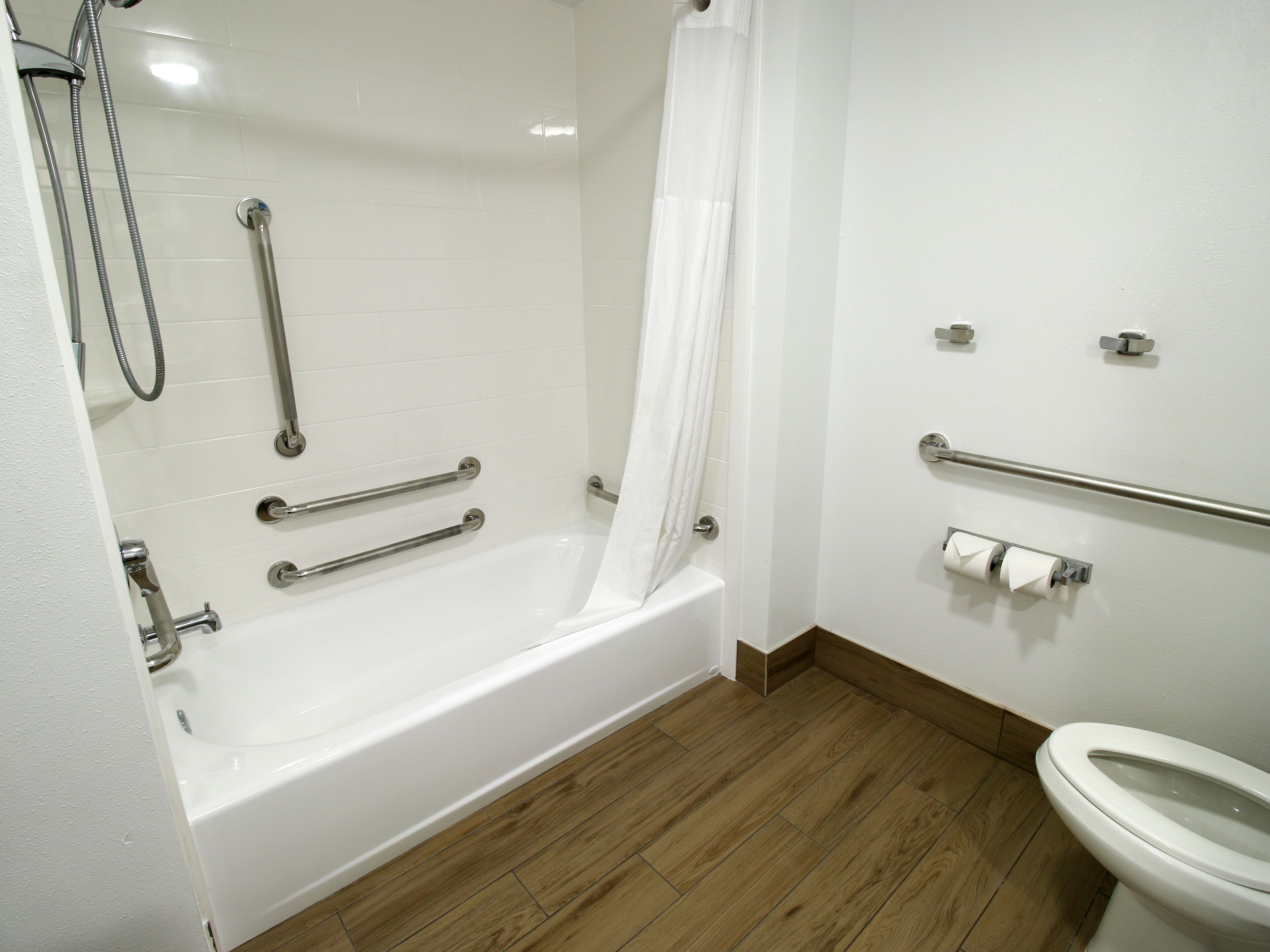 A bathroom with a white bathtub, wooden flooring, a toilet, and a shower with grab bars.